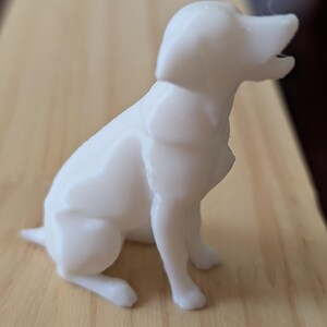 May include: A small, solid white figurine of a dog in a sitting position. The dog is depicted with its head turned slightly upward and its mouth open as if barking or panting. The surface of the figurine shows subtle horizontal 3D printed layers.