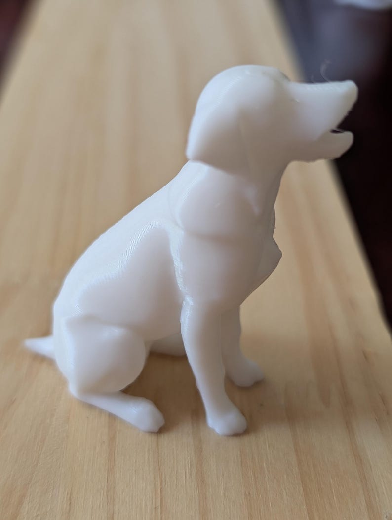 May include: A small, solid white figurine of a dog in a sitting position. The dog is depicted with its head turned slightly upward and its mouth open as if barking or panting. The surface of the figurine shows subtle horizontal 3D printed layers.