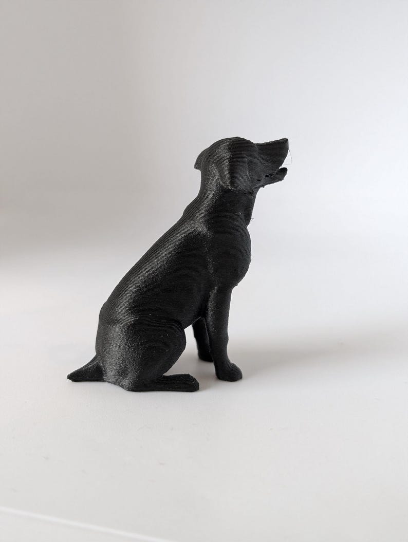 May include: A small, solid black figurine of a dog in a seated position. The sculpture features a textured surface with visible horizontal lines, a pointed snout, and ears that are folded down. The dog is facing toward the right side of the frame.