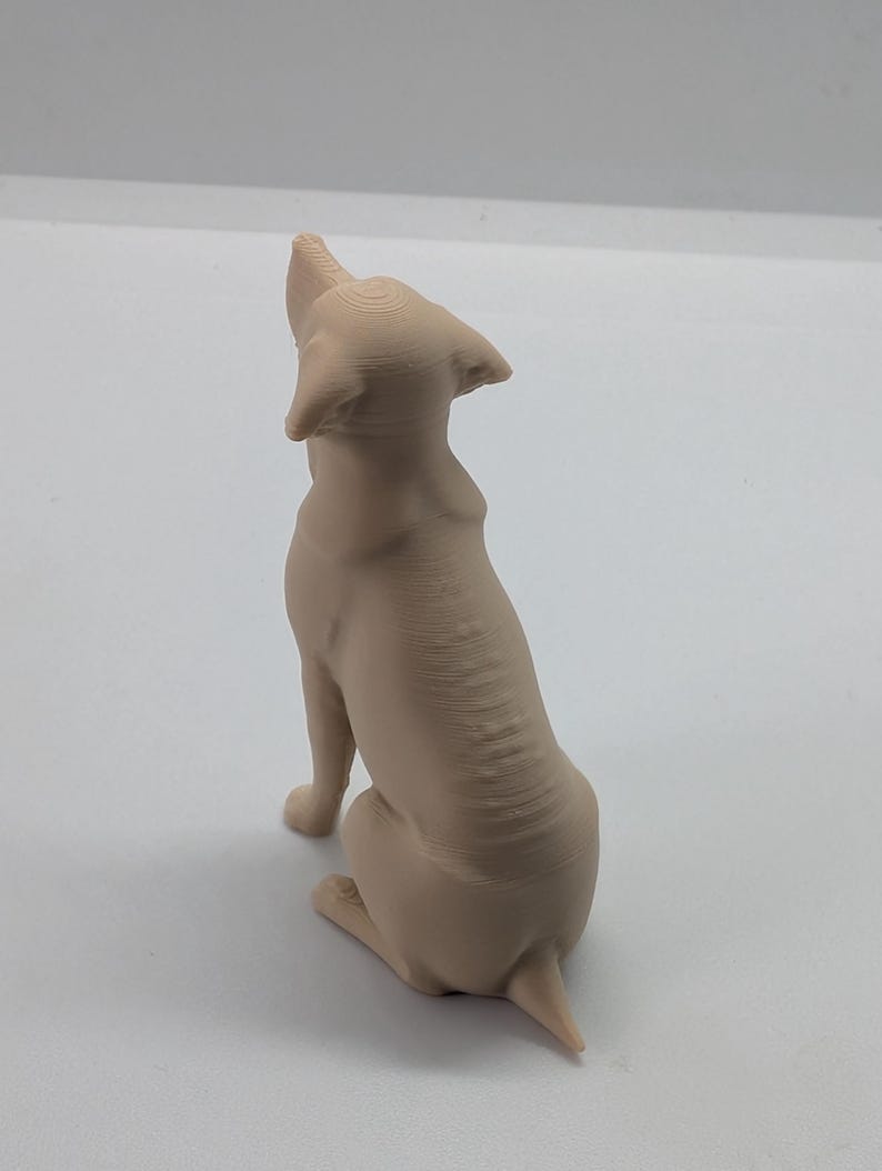 May include: A light beige, three-dimensional printed figurine of a dog sitting and looking upward. The sculpture features visible horizontal layer lines from the printing process, with pointed ears, a smooth back, and a short tail extending outward.