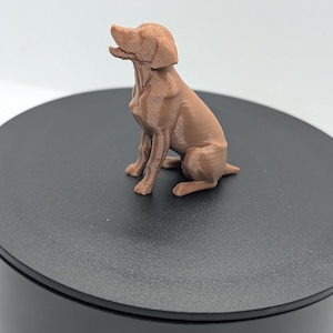 May include: A small, light brown, 3D-printed figurine of a dog in a seated position. The dog is depicted with its head tilted upward and mouth slightly open, showing visible horizontal layer lines from the printing process across its entire body.