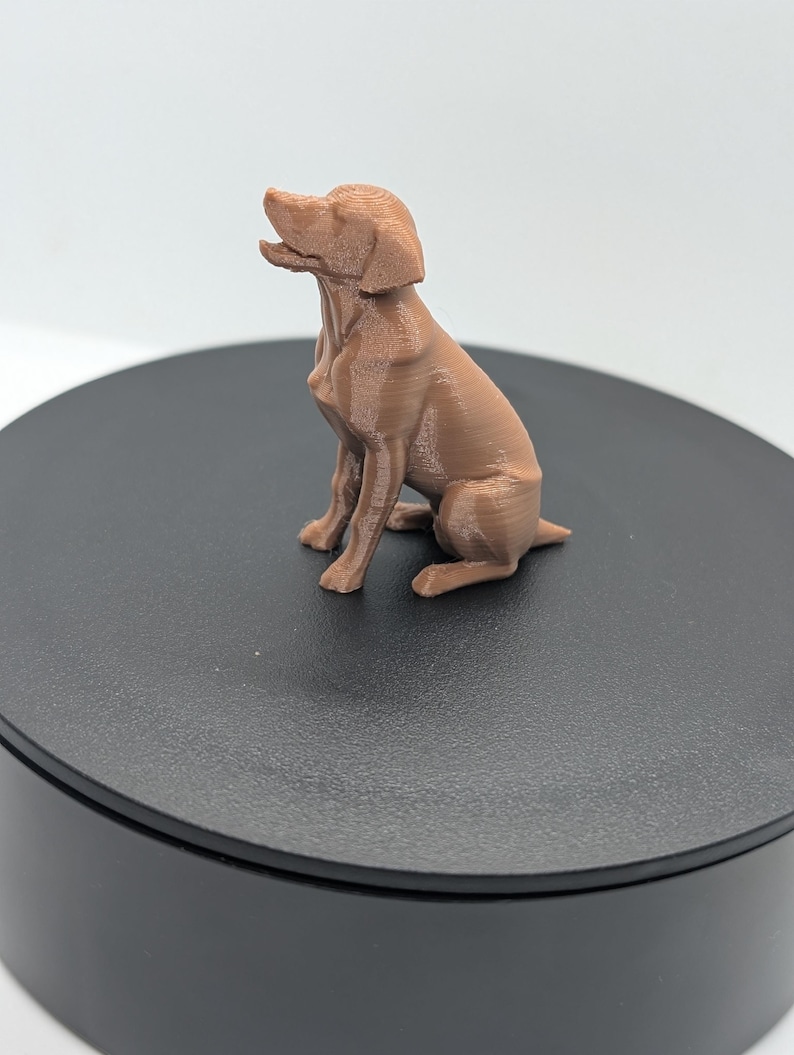 May include: A small, light brown, 3D-printed figurine of a dog in a seated position. The dog is depicted with its head tilted upward and mouth slightly open, showing visible horizontal layer lines from the printing process across its entire body.