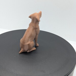 May include: A light brown, three-dimensional printed figurine of a dog in a seated position, viewed from the back. The dog has pointed ears and a short tail, with visible horizontal layer lines from the printing process on its surface.