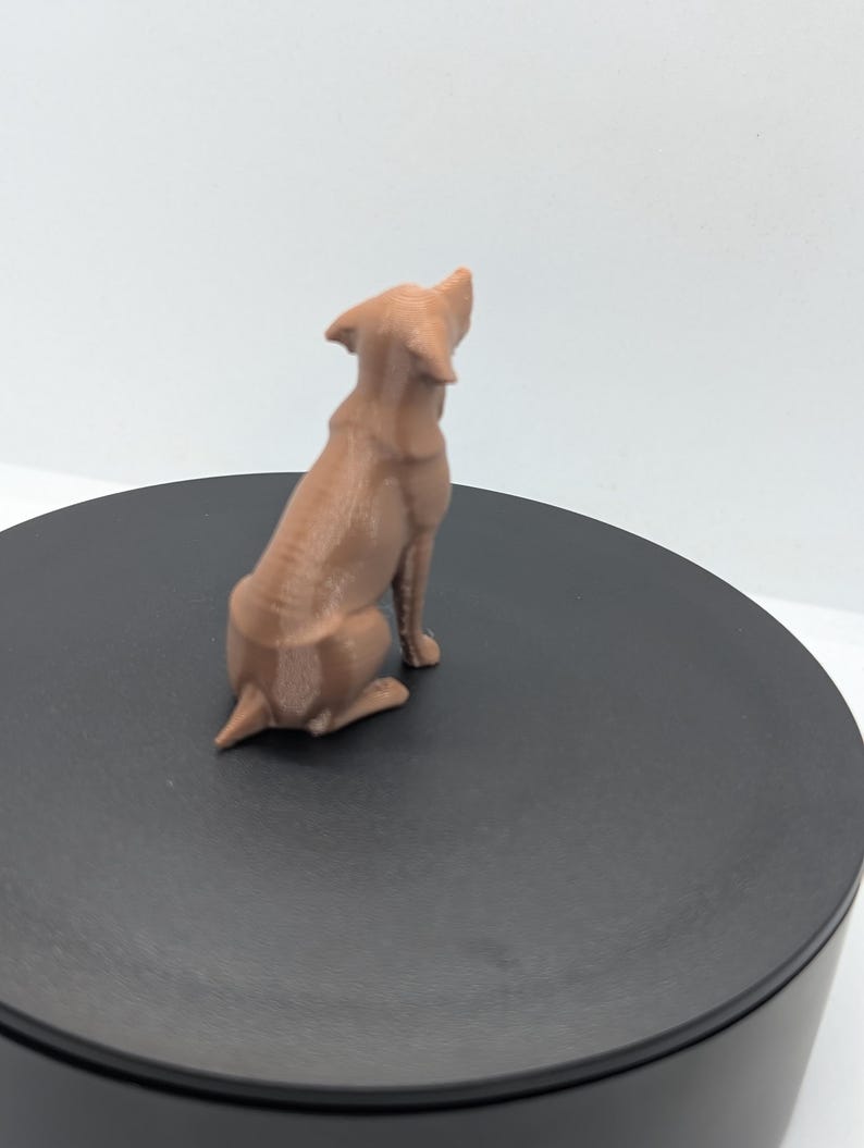 May include: A light brown, three-dimensional printed figurine of a dog in a seated position, viewed from the back. The dog has pointed ears and a short tail, with visible horizontal layer lines from the printing process on its surface.