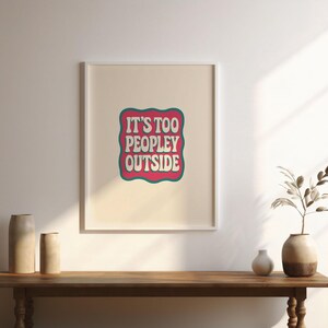 以下が含まれることがあります： レトロなフォントで「IT'S TOO PEOPLEY OUTSIDE」というテキストが書かれた額入りのプリント。テキストはピンクと白の配色で、ティール色のボーダーが付いています。プリントは、装飾的な花瓶が置かれた木製のテーブルに飾られています。