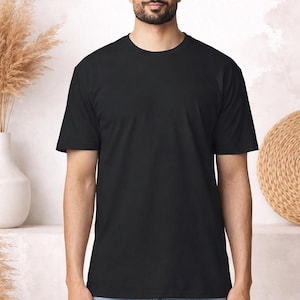 Peut inclure: Un t-shirt noir uni à col rond. Le t-shirt à manches courtes est fait d'une matière douce et est porté par une personne portant un jean bleu. L'arrière-plan présente un vase avec des plantes séchées et une décoration circulaire tissée.
