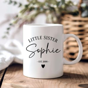 Könnte beinhalten: Ein weißer Keramikbecher mit schwarzem Aufdruck, der LITTLE SISTER liest, gefolgt von dem Namen Sophie in Schreibschrift, dem Text EST. 2008 und einem kleinen, ausgefüllten schwarzen Herzsymbol in der Mitte unter dem Design.