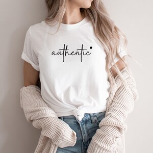 Könnte beinhalten: Weißes T-Shirt mit dem Wort "authentic" in handschriftlicher Schrift, mit einem kleinen Herz. Das Shirt wird mit blauen Jeans und einer cremefarbenen Strickjacke kombiniert. Der Look wirkt lässig und bequem.