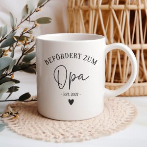 Könnte beinhalten: Eine weiße Keramiktasse mit dem schwarzen Schriftzug BEFÖRDERT ZUM Opa - EST. 2027 - und einem kleinen schwarzen Herzsymbol darunter. Die Tasse verfügt über einen Standardhenkel und einen glatten, zylindrischen Körper als Trinkgefäß.