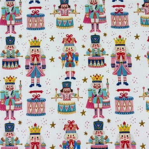 Könnte beinhalten: Ein weißer Stoff mit einem sich wiederholenden Muster aus Nussknackerfiguren und Trommeln in Rosa-, Blau- und Goldtönen. Die Nussknacker tragen Kronen und Uniformen, während die Trommeln mit Herzmotiven verziert sind und goldene Sterne.