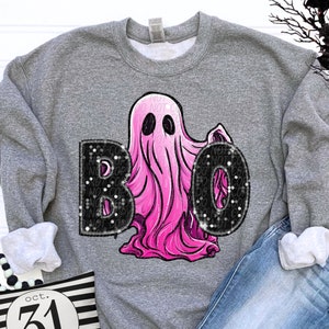 Op de afbeelding: Een grijs gem&ecirc;leerd sweatshirt met ronde hals en een afbeelding van een felroze geest tussen de zwarte letters B en O. De letters hebben een textuur met spikkels. Het sweatshirt wordt gecombineerd met een blauwe spijkerbroek.