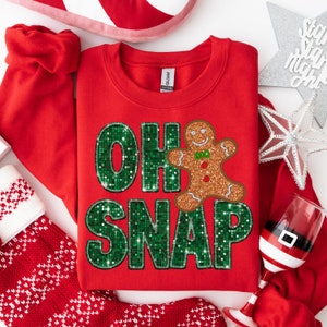 May include: Red sweatshirt with the phrase "OH SNAP" in green sequins and a gingerbread man graphic. The sweatshirt is surrounded by Christmas-themed decorations, including a stocking and a Santa-themed wine glass.