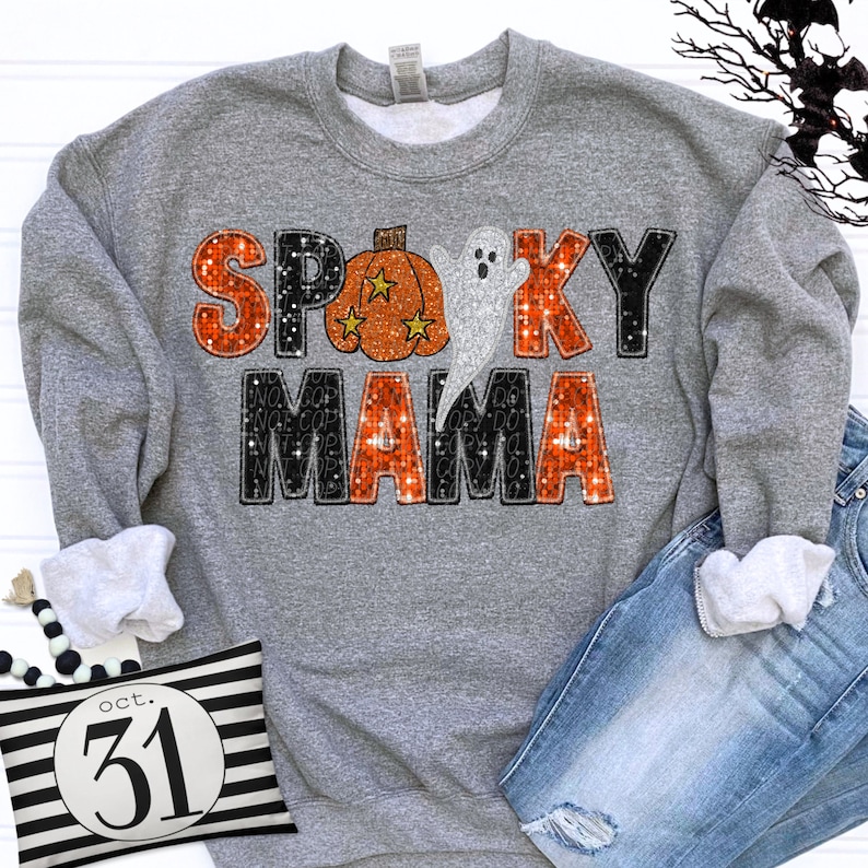 Puede incluir: Una sudadera gris jaspeado de cuello redondo con un estampado gr&aacute;fico que dice SPOOKY MAMA en letras brillantes naranjas y negras. La letra O se sustituye por una calabaza naranja con estrellas amarillas y un fantasma blanco sobre las letras.