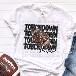 May include: White t-shirt with a black and silver glitter graphic of a football with the text "TOUCHDOWN Season" repeated multiple times.