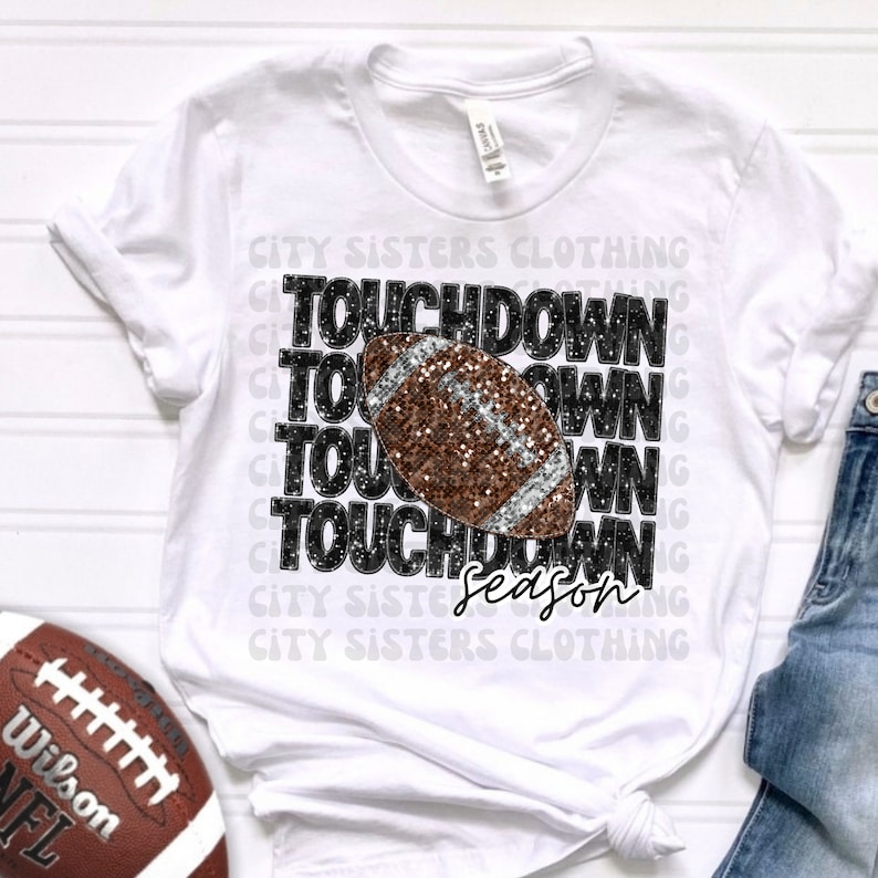 May include: White t-shirt with a black and silver glitter graphic of a football with the text "TOUCHDOWN Season" repeated multiple times.
