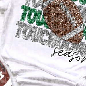 Op de afbeelding: Een wit T-shirt met een glinsterend zilver en groen "Touchdown Season" ontwerp. Een bruine en zilveren glitter voetbal is te zien in het ontwerp.