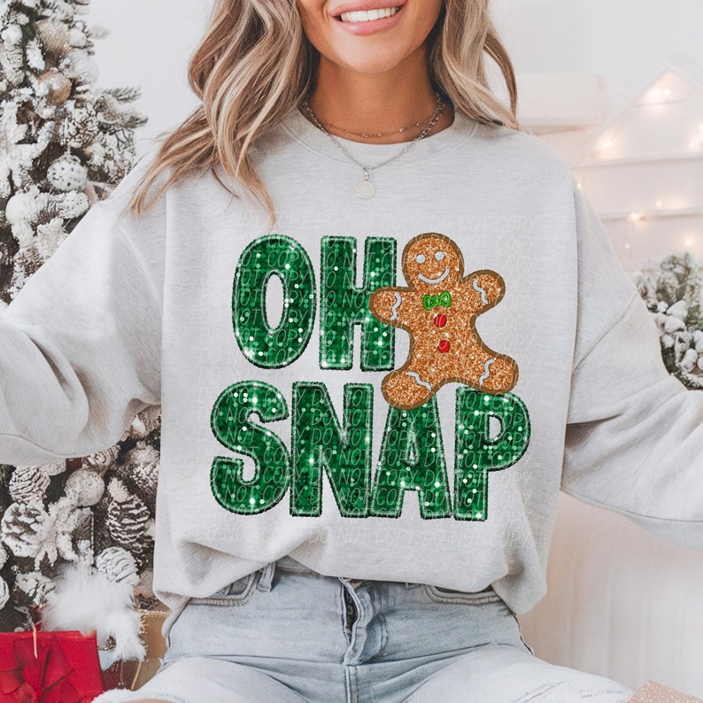 May include: A light gray sweatshirt with the phrase "OH SNAP" in green glitter letters and a gingerbread man graphic. The gingerbread man has gold glitter and red and green accents. The sweatshirt is a casual, festive design.