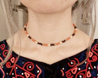 May include: A delicate choker necklace made with small brown, cream, and orange beads. The necklace is worn by a person with long brown hair.