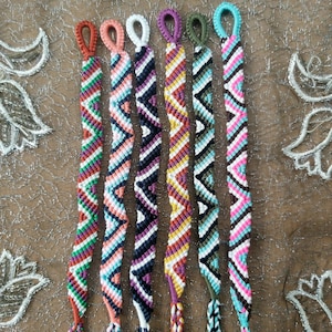 May include: Seven colorful friendship bracelets with geometric patterns in shades of green, orange, white, black, yellow, blue, and pink.