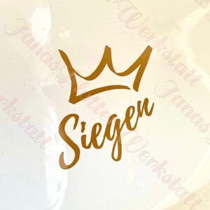 Könnte beinhalten: Goldene Krone mit dem Wort "Siegen" darunter in Kursivschrift.