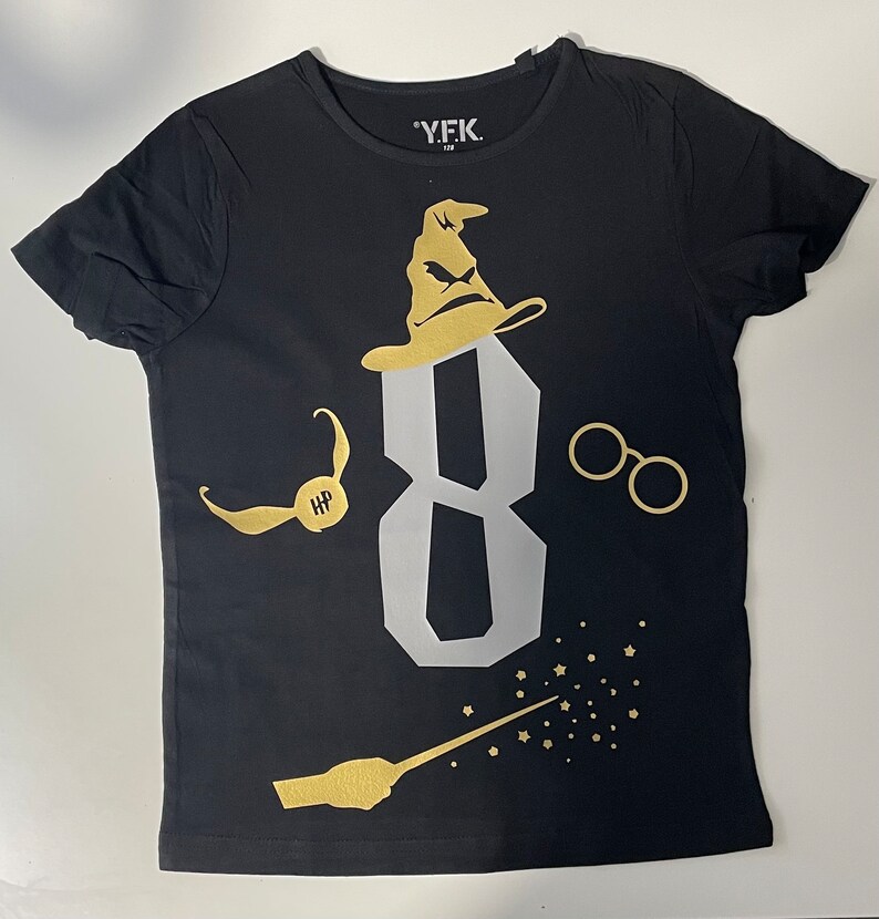 Pu&ograve; includere: Maglietta nera con una grafica dorata del numero 8, un Cappello Parlante, un paio di occhiali, un Boccino d'Oro e una bacchetta magica con stelle. Il testo "Y.F.K." &egrave; sulla maglietta.