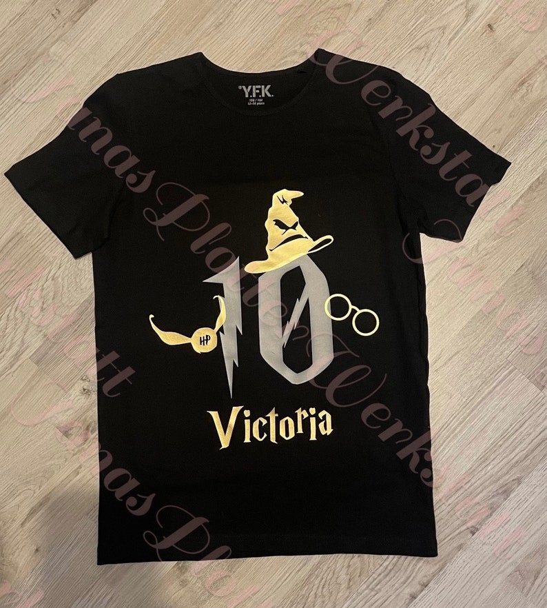 Pu&ograve; includere: Maglietta nera con una grafica dorata del numero 10, un cappello da mago, occhiali e un boccino d'oro. Il testo "Victoria" &egrave; sotto il numero.