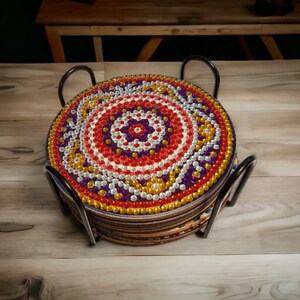 Op de afbeelding: Een set van zes kleurrijke onderzetters met een geometrisch patroon, bestaande uit rode, gele, paarse en witte kralen. De onderzetters worden bij elkaar gehouden door een zwarte metalen standaard.