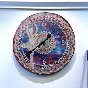 May include: A round clock with a ballerina silhouette and a galaxy-themed background. The clock face is decorated with rhinestones and pearls. The clock hands are black and point to the numbers 12 and 3.