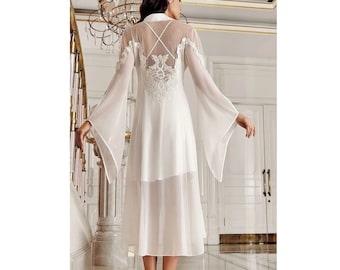 May include: A white nightgown with long, sheer bell sleeves and a decorative lace back. The gown is floor-length and has a layered skirt and a high collar. The back of the gown has a criss-cross design.