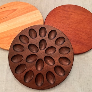 May include: Three round wooden serving trays. One tray is made of light wood with a natural finish. Another tray is made of dark wood with a natural finish. The third tray is made of dark wood with a natural finish and has 18 oval-shaped indentations.