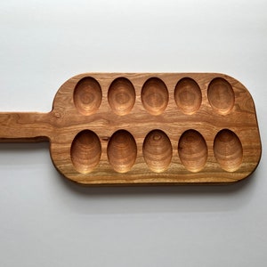 May include: A wooden serving tray with a handle, featuring 12 oval-shaped indentations for holding eggs. The tray is made of light brown wood with a natural grain pattern.