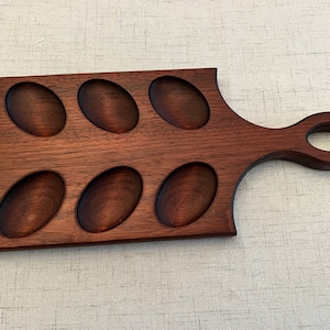 May include: A wooden egg tray with a handle. The tray has six oval-shaped indentations for holding eggs. The tray is made of dark brown wood and has a natural finish.