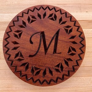 Könnte beinhalten: Ein runder Holz-Untersetzer mit einem geschnitzten geometrischen Muster und dem Buchstaben "M" in der Mitte.