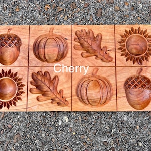 May include: A rectangular wooden plaque with eight carved autumn-themed designs. The designs include acorns, pumpkins, oak leaves, and sunflowers. The word "Cherry" is written across the center. The wood has a warm, reddish-brown color.