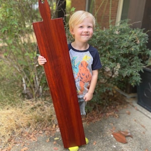 May include: A young child holds a large, rectangular wooden cutting board with a red-brown stain. The board has a decorative handle at the top.