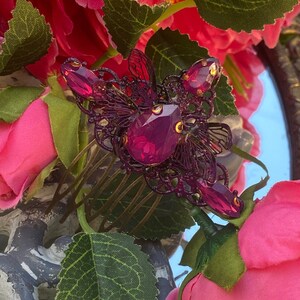 May include: A purple butterfly hair comb with a large, dark pink rhinestone in the center. The comb is surrounded by pink roses.