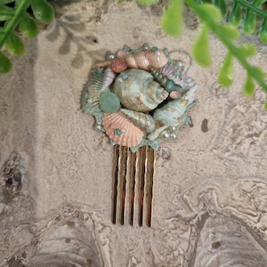Puede incluir: Un peine de pelo dorado adornado con un grupo de conchas y perlas. Las conchas son en tonos de rosa, verde y blanco. El peine es un accesorio encantador para una boda en la playa o una fiesta de verano.
