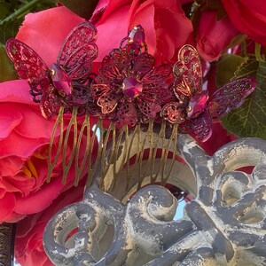 May include: A gold hair comb with a decorative design of three pink butterflies with rhinestones. The comb is set against a background of pink roses.