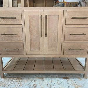 May include: A light-colored wooden bathroom vanity with six drawers and two cabinet doors. The drawers and doors have bronze-colored hardware. The vanity has an open shelf at the bottom and is constructed with a natural wood finish.