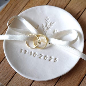 Könnte beinhalten: Eine weiße Keramik-Ring Schale mit einem zarten floralen Design und der eingravierten Datum 17-10-2025. Zwei goldene Eheringe liegen auf der Schale und sind mit einem weißen Satinband zusammengebunden.