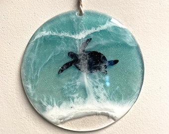 May include: A round, clear resin ornament with a blue and white wave design and a black silhouette of a sea turtle swimming in the water.