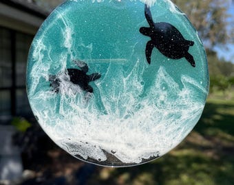 May include: Round teal and white resin ornament with two black turtle silhouettes. The ornament is hanging from a string.