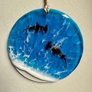 May include: A round, ocean-themed resin art piece featuring two black hammerhead sharks swimming in a blue and white ocean. The artwork is suspended by a natural fiber cord.