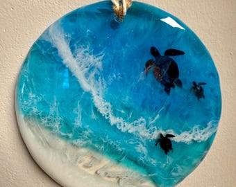 May include: Round resin art piece depicting a beach scene with three black sea turtles swimming in turquoise water. The artwork features white foamy waves and a sandy shore. A natural twine loop is attached for hanging.