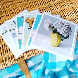 May include: Five blank greeting cards with floral designs. The cards are arranged in a fan shape on a wicker basket with a blue and white checkered fabric underneath.