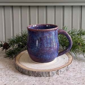 May include: A ceramic mug with a rounded body and a curved handle, featuring a glossy glaze in deep shades of purple and blue with subtle mottled textures. The mug has a smooth, rounded rim and a solid base, showcasing a rich, multi-tonal finish.