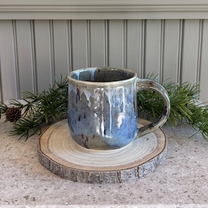 May include: A ceramic mug with a reactive glaze featuring shades of blue, gray, and white with dark brown speckles. The mug has a rounded body, a sturdy handle, and a smooth rim, showcasing a dripped glaze effect that creates a unique pattern.