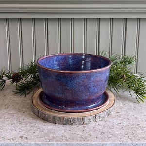 May include: A round ceramic bowl with a glossy finish featuring a deep blue and purple mottled glaze. The bowl sits on a matching circular saucer, which is placed on a thick, natural wood slice with visible bark edges and green pine needles.