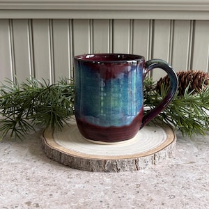 May include: A ceramic mug featuring a glossy glaze that transitions from a deep burgundy at the base and rim to a vibrant teal and blue in the center. The mug has a curved handle and a smooth, rounded body, showcasing a rich, multi-toned finish.