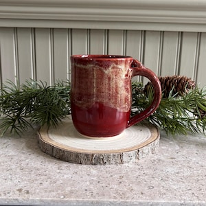 May include: A ceramic mug with a deep red base and a textured, lighter cream-colored glaze dripping down from the rim. The mug features a sturdy handle on the right side and is positioned on a circular wooden slice with visible bark edges.